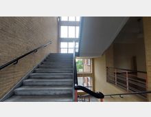 Treppenhaus in einem Gebäude mit Wänden aus gelben Ziegelsteinen, einem Treppengeländer aus schwarzem Metall und großen Fenstern, die viel Tageslicht hereinlassen. Die Fenster bieten einen Blick auf eine Außenszene mit Bäumen und anderen Gebäuden.