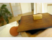 Holzkiste mit einem Griff auf dem Deckel im Vordergrund, daneben ein orangefarbener Gegenstand auf einem weißen Handtuch; im Hintergrund unscharf ein heller Raum mit Holzboden, weißen Wänden und einer Zimmerpflanze.