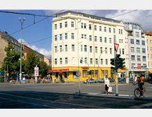 Straßenkreuzung in einer Stadt mit einer gelben, mehrstöckigen Gebäudefassade, in der sich eine Apotheke befindet. Im Vordergrund sind eine Ampel, ein Fahrradfahrer und eine Fußgängerin zu sehen; im Hintergrund verläuft ein Gehweg mit Bäumen und weiteren Gebäuden.