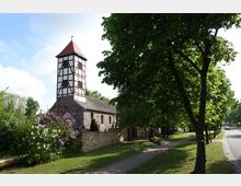 Fachwerkkirche mit quadratischem, rot gedecktem Turm und Mauern aus Feldsteinen, umgeben von Blumengewächsen und einer niedrigen Steinmauer, neben einem Gehweg mit Bäumen in einer grünen Umgebung.