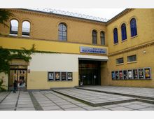 Backsteinfassade eines Gebäudes mit gelben Akzenten, das zum "Kino in der Kulturbrauerei" gehört. Der Eingang ist durch Glas- und Metalltüren gekennzeichnet, daneben mehrere Schaukästen mit Filmplakaten; ein gepflasterter Vorplatz und ein Durchgang mit Efeubewuchs runden die Szenerie ab.