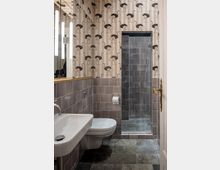 Kleines Badezimmer mit einer Wandtapete mit Straußenmotiv, beigefarbenen und grauen Fliesen an den Wänden, einem weißen Waschbecken, einer Toilette und einer verglasten Dusche. Der Boden ist mit dunkelgrauen Fliesen ausgelegt.