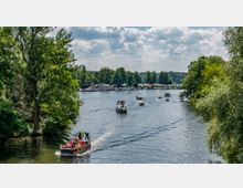 Flusslandschaft mit mehreren motorisierten und kleinen Booten auf dem Wasser, umgeben von dichtem Grün und Bäumen. Im Hintergrund sind eine Uferpromenade und geparkte Boote sichtbar, unter einem bewölkten Himmel.