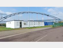 Ein flaches, weißes Bürogebäude mit der Beschriftung „HLB-Hafenmeister“ steht vor einer großen, modernen Brücke mit blauem Stahlbogen und roten Aufhängeseilen. Im Vordergrund ist eine gepflasterte Straße, im Hintergrund sind Strommasten und Grünflächen zu sehen.