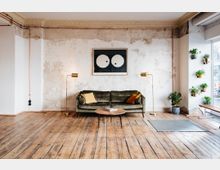 Wohnraum mit rustikalem Holzfußboden, einer grünen Samtcouch mit gelben und orangefarbenen Kissen, flankiert von zwei goldenen Stehlampen. Die Wand im Hintergrund weist eine abgeblätterte Textur auf, dekoriert mit einem minimalistischen Kunstwerk, und auf der rechten Seite hängen Pflanzen in Töpfen.