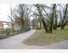 Ein parkähnlicher Weg mit kahlen Bäumen und vereinzeltem Grasbewuchs, der an einem Maschendrahtzaun mit angrenzendem Spielplatz vorbeiführt. Im Hintergrund sind Backsteingebäude und weitere Vegetation zu sehen.