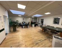 Großraumbüro mit Holzboden, hellem Deckenlicht und mehreren Arbeitsplätzen. Links befinden sich Pflanzen und ein Glastrenner mit der Aufschrift "Vorstufe", rechts ein Konferenztisch mit Stühlen.