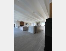 Modernes Wohnzimmer mit hellen Wänden, einer Decke mit sichtbaren Balken und großen Fliesenböden. Im Raum befinden sich ein beiges Sofa, ein Marmortresen, eine Fernsehecke und große Fenster mit weißen Vorhängen, die viel Tageslicht hereinlassen.