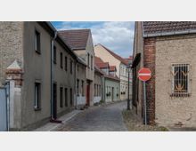 Schmale Kopfsteinpflastergasse mit angrenzenden Wohnhäusern aus Putz und Backstein, teilweise mit Blumenkästen an den Fenstern; im Vordergrund ein „Einfahrt verboten“-Schild und eine alte Laterne.