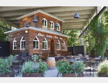 Holzgebäude mit der Aufschrift "Oberhafen-Kantine" unter einem offenen Betondach, umgeben von zahlreichen Tischen und Stühlen im Außenbereich. Im Vordergrund befinden sich Pflanzkübel mit grünen Pflanzen.