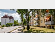 Straßenszene mit Wohngebäuden: Rechts ein mehrstöckiges gelbes Wohnhaus mit Balkonen, geschmückt mit Blumen und Markisen, links ein weiteres Wohnhaus mit Satteldach. Dazwischen verläuft eine schmale, asphaltierte Straße, gesäumt von Bäumen und einem Gehweg.