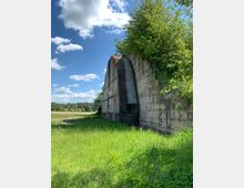 Eine alte Betonmauer mit gewölbter Struktur, teils überwuchert von Bäumen und Sträuchern, steht am Rand einer grünen Wiese unter einem Himmel mit vereinzelten Wolken. Im Hintergrund ist Wald zu erkennen.