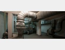 Industrie- oder Technikraum mit großen Lüftungsrohren und Ventilationsanlagen, die an der Decke und den Wänden entlang verlaufen. Der Raum hat unverputzte Betonwände und eine karge Beleuchtung.