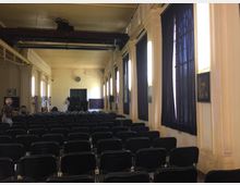 Ein Veranstaltungsraum mit Reihen schwarzer Stühle, hohen cremefarbenen Wänden, großen Fenstern mit dunkelblauen Vorhängen und einer sichtbaren Stahlträger-Konstruktion an der Decke. Im Hintergrund befindet sich eine Uhr sowie technische Geräte an der Wand.