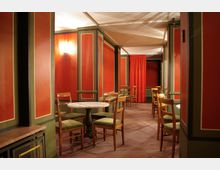 Ein gemütlicher Innenraum mit orangefarbenen Wänden und grünen Holzverkleidungen, ausgestattet mit mehreren runden Tischen, Holzstühlen mit grünen Polstern und einer roten Vorhangabtrennung im Hintergrund. Die Beleuchtung erfolgt durch Wandleuchten, die warmes Licht spenden.