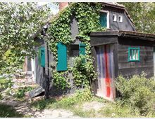 Schwarzes Holzhaus mit grünen Fensterläden, bewachsen mit Efeu. Im Vordergrund ein blühender Fliederbaum, rechts ein Eingang mit bunter Kunststofflamellen-Vorhang und links ein kleiner Sitzbereich mit rotem Gartentisch und Stühlen.