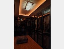 Ein begehbarer Kleiderschrank mit Holzfußboden, Kleiderschränken mit Glastüren und integrierter Beleuchtung. Eine Kristall-Deckenleuchte und warme Lichtakzente erzeugen eine elegante Atmosphäre; in der Mitte steht ein gepolsterter Hocker.