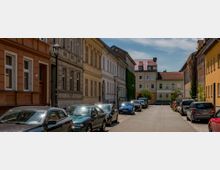 Eine schmale, gepflasterte Stadtstraße mit parkenden Autos auf beiden Seiten, gesäumt von mehrstöckigen Wohngebäuden mit farbigen und verzierten Fassaden; im Hintergrund ein modernes Gebäude mit einem roten Ziegeldach.