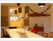 Esszimmer mit einem langen weißen Tisch und verschiedenen Stühlen, dekoriert mit hängenden Lampen und einer Geburtstagsgirlande an der Wand. Im Hintergrund ist eine helle Küche mit Holzschränken sichtbar, und an der Wand befindet sich ein Holzregal mit Dekorationen.