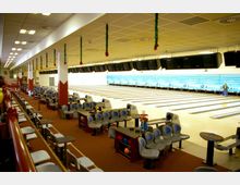 Innenraum einer Bowlinghalle mit mehreren Bahnen, Sitzbereichen und Tischen. An der Decke hängen dekorative Girlanden, und oberhalb der Bahnen sind Monitore angebracht; im Hintergrund ist eine blaue Wand sichtbar.