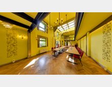 Gelb gestrichener Tagungsraum mit hohen Decken, Holzparkett und großen Fenstern, die Tageslicht hereinlassen. Ein langer Tisch mit roten Polsterstühlen und dekorativen Elementen an den Wänden, darunter filigrane Muster und Kronleuchter, prägt das moderne, elegante Interieur.