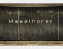 Wand eines U-Bahnhofs mit dem Schriftzug „Haselhorst“ in großen, silbernen Buchstaben auf einer dunklen, metallischen Oberfläche mit sichtbaren Alterungsspuren. Am unteren Rand ist ein Boden mit beigen und weißen Fliesen zu sehen.