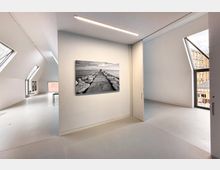 Heller, minimalistischer Innenraum mit schrägen Dachfenstern, weißen Wänden und glattem Boden, an dem eine Schwarz-Weiß-Fotografie eines Stegs über dem Meer hängt. Im Hintergrund befinden sich Fenster mit Blick auf umliegende Gebäude.