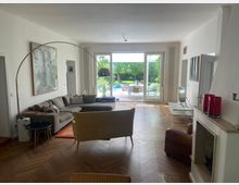 Wohnzimmer mit Parkettboden, einem großen Ecksofa, einem gelben Sessel und einem modernen Kamin. Große Glas-Schiebetüren führen zu einem Garten mit Pool und Terrasse im Hintergrund.