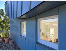 Außenansicht einer Gebäudefassade mit blauer Metallverkleidung und zwei weißen, rechteckigen Fenstern. Im Vordergrund stehen mehrere Blumentöpfe auf einem mit Kies bedeckten Boden, umgeben von grüner Vegetation.