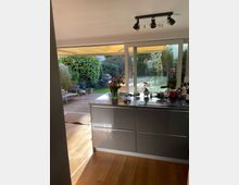 Wohnküche mit glänzend grauer Kücheninsel, Holzfußboden und großen Glasfronten, die auf eine Holzterrasse mit Blick in einen grünen Garten führen. Auf der Kücheninsel stehen Blumen und Küchenutensilien, während im Garten Pflanzen und Gartenmöbel sichtbar sind.