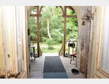 Blick aus einer Holzveranda mit Rundbogen und Säulen ins Grüne, umgeben von Bäumen. Auf der Veranda befinden sich ein Tisch mit Stuhl, eine Fußmatte und dekorative Gegenstände; im Hintergrund ist ein geparktes Auto sichtbar.