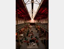 Innenansicht einer renovierten Industriehalle mit hohen Decken und einem Glasdach, das Tageslicht hereinlässt. Der Raum ist mit modernen Sitzgruppen in verschiedenen Farben, kleinen Tischen und Pflanzen dekoriert, während die Wände und tragenden Stahlträger die ursprüngliche Bausubstanz zeigen.