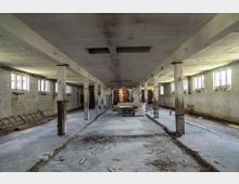 Großzügiger, verlassener Innenraum eines alten industriellen oder landwirtschaftlichen Gebäudes mit Betonboden, Pfeilern, heruntergekommenen Wänden und mehreren Fenstern, die Tageslicht hereinlassen. In der Mitte des Raumes stehen ein Sofa, ein Tisch und ein paar Stühle, umgeben von Spuren von Verfall und verstreuten Gegenständen.