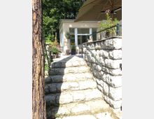 Steintreppe, die zu einer erhöhten Terrasse mit Geländer führt, auf der Pflanzen in Töpfen, ein Tisch mit Stühlen und ein großer Sonnenschirm stehen; im Hintergrund ein kleines Gebäude mit cremefarbener Fassade und großen Fenstern, umgeben von Bäumen.