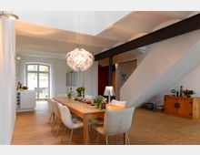 Helles, modernes Esszimmer mit großem Holztisch und acht gepolsterten Stühlen, einer auffälligen Deckenleuchte und Holzboden; im Hintergrund große Fenster und Treppe, die ins obere Geschoss führt.