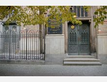 Historische Gebäudefassade mit verzierten Metalltüren, eingefasst von steinernen Säulen und einem Metallzaun im Vordergrund. An der Wand befindet sich eine Gedenktafel mit hebräischem Stern und Text, umgeben von gelb-grünen Ästen eines Baumes.