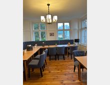 Ein heller Innenraum eines Cafés oder Restaurants mit Holzboden, großen weißen Sprossenfenstern und einem zentralen, modernen Kronleuchter. Der Raum ist mit Holztischen und gepolsterten grauen Stühlen ausgestattet, teilweise mit blauer Wandpolsterung im Hintergrund.