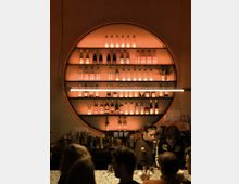 Eine Bar mit einer halbkreisförmigen, beleuchteten Rückwand, auf der Flaschen in mehreren Reihen stehen. Im Vordergrund befinden sich eine Theke mit Getränken und Gästen sowie zwei Personen, die hinter der Bar arbeiten.