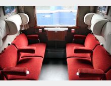 Innenraum eines Zugabteils mit zwei gegenüberliegenden Sitzbänken in rotem Polsterstoff. In der Mitte befindet sich ein kleines klappbares Holztischchen unter einem Fenster, durch das ein blauer Waggon sichtbar ist.