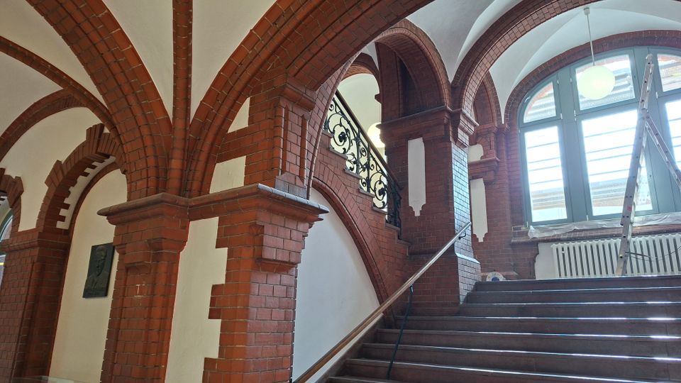 Innenraum eines historischen Treppenhauses mit roten Backsteinbögen und weißem Gewölbe, einer breiten Steintreppe mit Geländer sowie dekorativem Schmiedeeisengitter. Großes Rundbogenfenster mit Heizkörper darunter; eine Leiter steht am Fensterbereich.