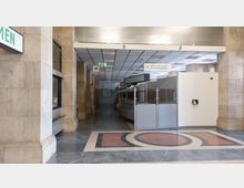 Eingangsbereich eines öffentlichen Gebäudes mit beigefarbenen Steinwänden, Glas-Trennwänden und einem Boden aus poliertem Stein mit geometrischen Mustern. Im Hintergrund befinden sich Schalter und Schilder, darunter ein "Bitte nicht rauchen"-Hinweis und ein grünes Fluchtwegsymbol.