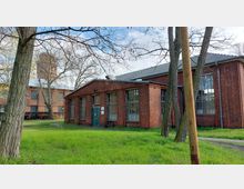 Backsteingebäude mit großen Fenstern und einem Satteldach, umgeben von Gras und Bäumen; im Hintergrund ist ein weiteres Backsteingebäude mit einem Turm zu sehen. Der Bereich wirkt wie ein historisches Industrie- oder Werksgelände.