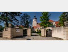 Eingang zu einer historischen Kirche mit hellen Steinmauern, einem geschwungenen Torbogen und einem Weg, der zum Gebäude führt. Die Kirche hat ein rotes Ziegeldach und einen hohen Turm mit spitzer Turmhaube, umgeben von Bäumen und grünem Rasen.