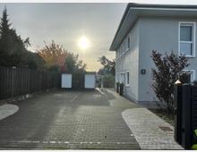 Wohngebiet mit einem zweistöckigen hellgrauen Gebäude auf der rechten Seite, gepflastertem Hof und zwei Garagen am Ende des Weges. Im Hintergrund ist ein Baum mit Herbstlaub zu sehen, und die Abendsonne scheint durch einen leicht bewölkten Himmel.