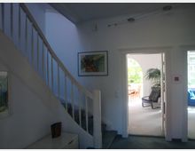 Wohnbereich in einem Haus mit weiß gestrichener Treppe, Bildern an den Wänden und einem angrenzenden Raum mit großen Fenstern, einem Esstisch aus Holz und einer schwarzen Sessel. Im Hintergrund ist eine Palme erkennbar.