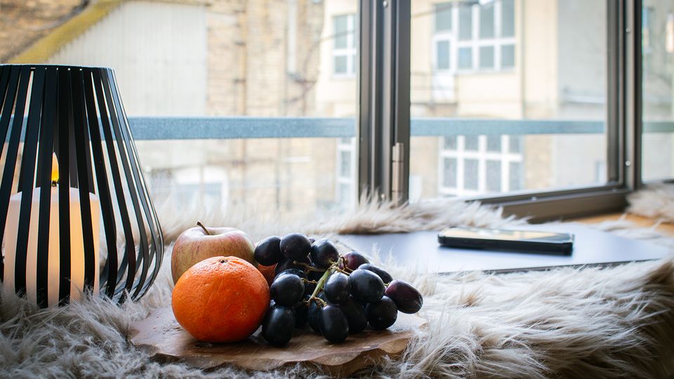 Fensterbank in einem Innenraum mit großer Fensterfront; darauf stehen eine schwarze Laterne mit Kerze, ein Holzbrett mit Obst sowie ein Laptop auf einem flauschigen Fell. Durch das Fenster sind gegenüberliegende Gebäude mit Ziegel- und Putzfassaden und mehreren Fenstern zu sehen.
