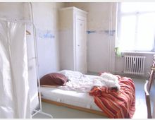 Ein einfach eingerichtetes Schlafzimmer mit einer Matratze auf dem Boden, darauf unordentliche Bettwäsche und eine gestreifte Decke. Im Hintergrund befinden sich ein kleiner weißer Schrank, ein Fenster mit Heizkörper darunter und eine Pflanze auf der Fensterbank.