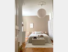 Helles Schlafzimmer mit beiger Wandfarbe, einem Doppelbett mit weißer Bettwäsche und dekorativen Kissen. Im Raum befinden sich ein kleiner Hocker mit Dekoration, ein hängender runder Papierlampenschirm und weiße Schränke; der Boden besteht aus Holzdielen.