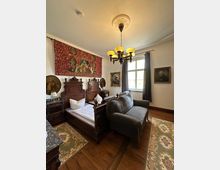 Ein historisch eingerichtetes Schlafzimmer mit einem Einzelbett aus dunklem Holz, einem gepolsterten Sofa im antiken Stil und Teppichen auf einem Holzboden. An der Wand hängen ein roter Wandteppich mit mittelalterlichen Motiven und mehrere Gemälde; ein Kronleuchter mit gelbem Licht hängt von der Decke, und ein Fenster mit Vorhängen erhellt den Raum.