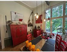 Esszimmer mit großer Fensterfront und Blick ins Grüne, eingerichtet mit roten Holzmöbeln, darunter ein rustikaler Schrank und Stühle. Der Raum ist mit warmem Licht, Kerzen und dekorativen Kunstgegenständen gestaltet.